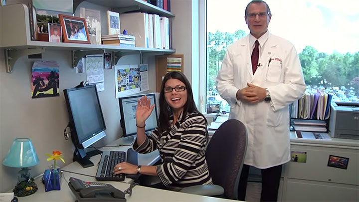 A physician speaking to the camera whilst standing next to a person sitting at their office computer desk